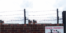 brick wall with barbed wire on top and a sign that has been grafittied to read Private Property is Theft